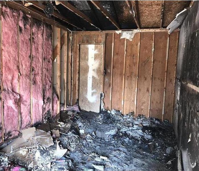 Room with drywall burned and fallen to floor and soot-filled insulation on walls.  Also black smoke damage on ceiling joists 
